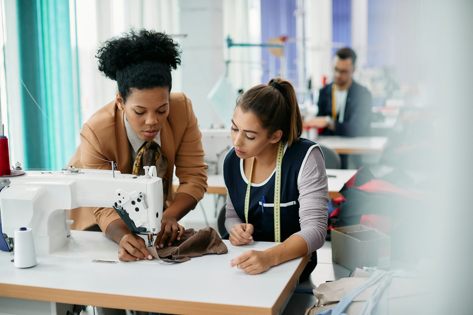 Black clothing designer assisting young seamstress with sewing in a textile workshop.